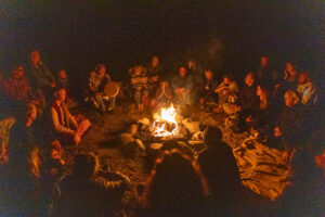 A group of over two dozen people in the dark night sitting by a blazing campfire.