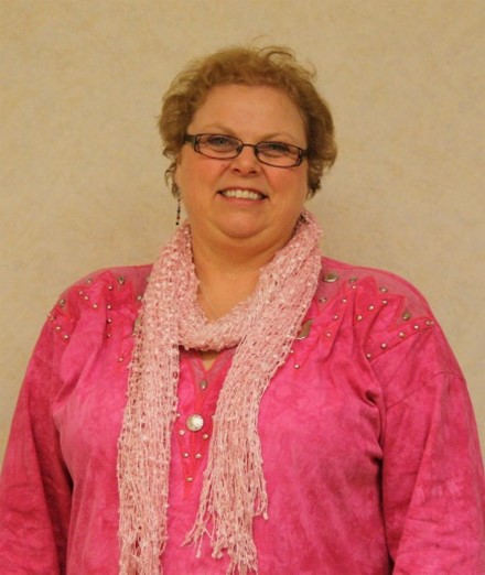 A white woman with short brown hair smiles at the camera in front of a tan background. She is wearing a bright pink shirt and a light pink scarf and glasses.
