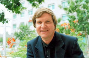 A smiling white man with blond hair is sitting in front of flowers and trees. He is wearing a black button up and black blazer.