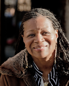 A Black woman with long braids smiles at the camera. She is wearing long earrings, a striped shirt, and a tan jacket with a fur collar.