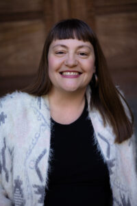 A white woman with long brown hair wearing a white sweater smiles at the camera.