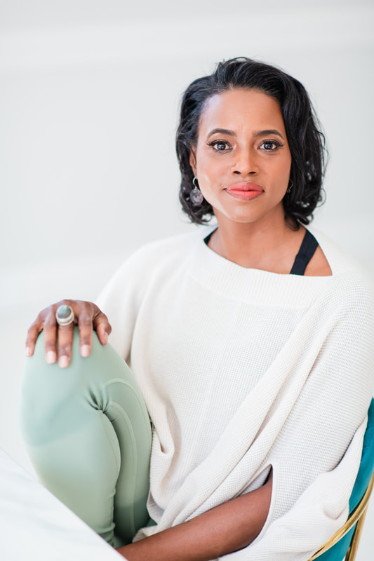 A Black woman with medium length dark hair wearing a white shirt and light green pants in front of a light background.