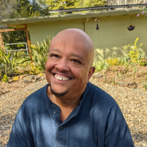 A smiling, bald, Black man in a blue shirt in front of a garden.