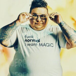 A man with curly hair has both hands putting on his glasses while looking straight ahead. He is wearing a tshirt that says "Fuck normal I want magic."