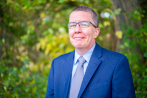 A white man with short hair and glasses is grinning at the camera. He wears a blue suit in front of a forested background.