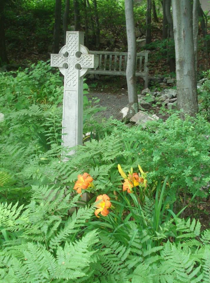 A stone cross in a lush green garden with daffodils in the foreground.