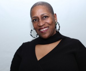 A Black woman with short hair and wearing earrings in front of a white background.