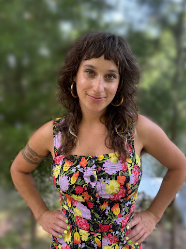 A headshot of a light-skinned person with long dark hair wearing a colorful floral top.