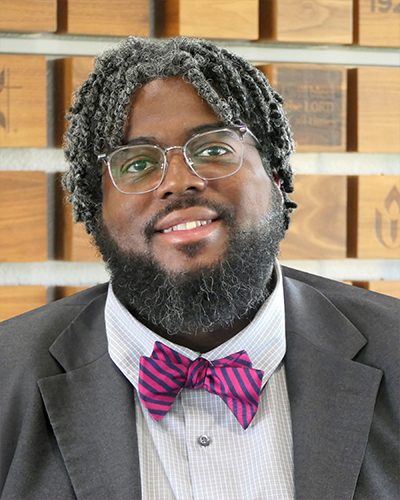 A smiling Black man with a beard, medium locs, and glasses wearing a white shirt, striped bowtie, and suit jacket.