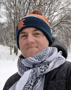 A grinning white man is bundled in front of a snowy background. He is wearing a beanie with a sports logo and a keffiyeh scarf.