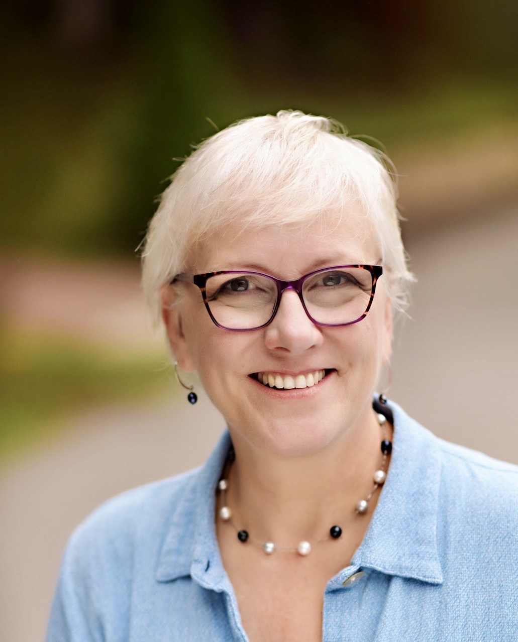 A headshot of a white woman with medium-length blonde hair in glasses and a blue shirt.