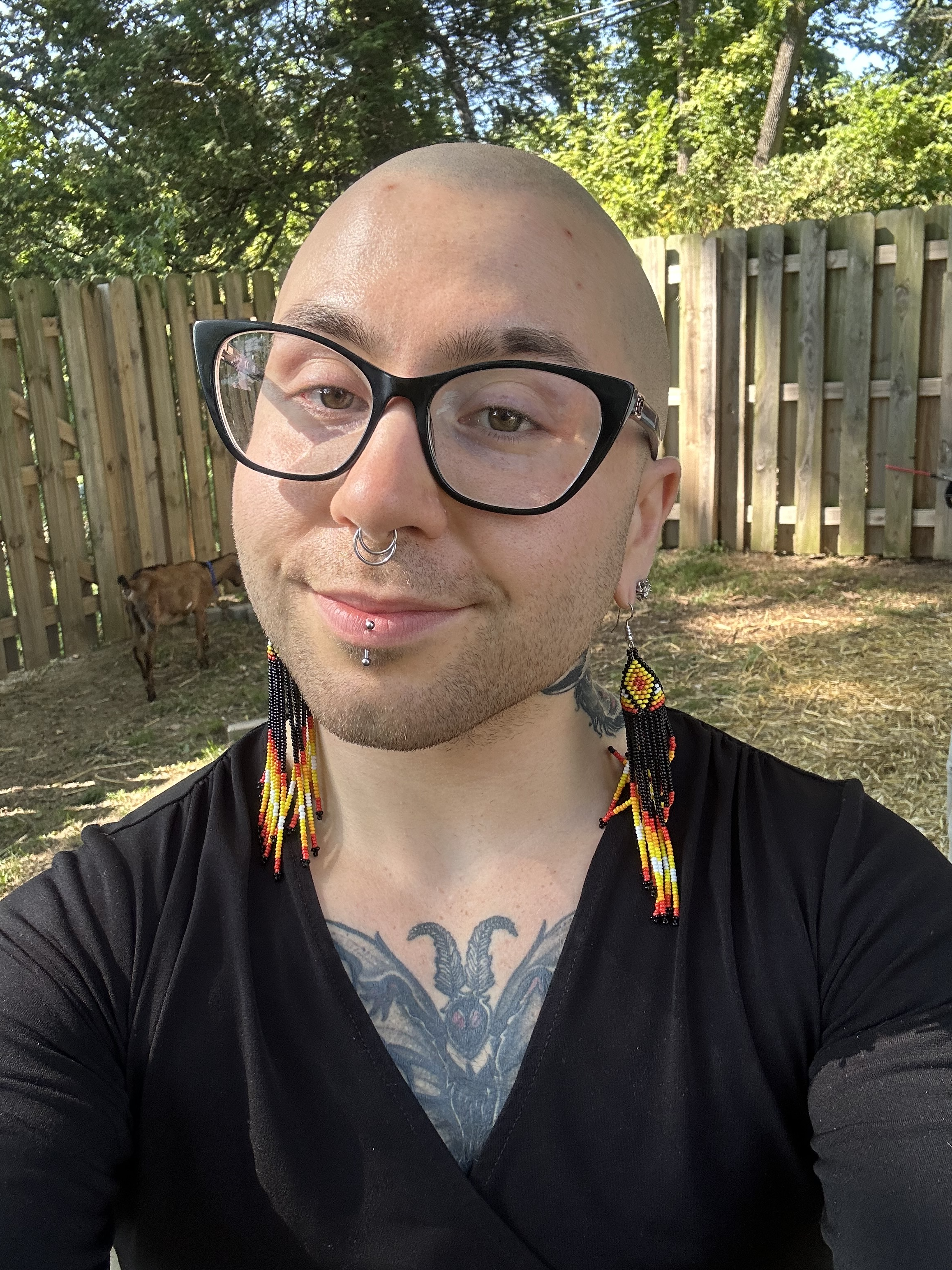 A light-skinned person with a shaved head, glasses, and long earrings in front of a wooden fence.