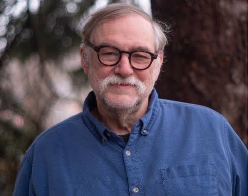 A headshot of a white man with a white mustache and glasses in a blue shirt.