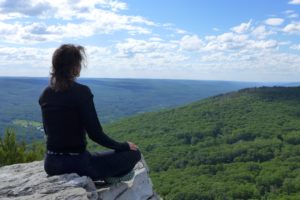 Woman in black sits cross legged in a meditation pose at the edge of a cliff