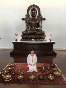 a woman in white sits before a giant copper Buddha, ringed by singing bowls