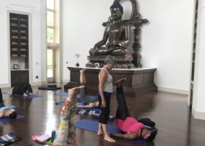 A yoga instructor helps a participant in a yoga class with a large Buddha statue in the back