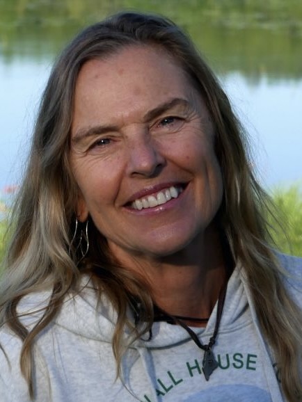 A smiling woman with long blond hair standing in front of a lake