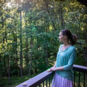 A woman on retreat standing on a deck and gazing out to the lush, green forest