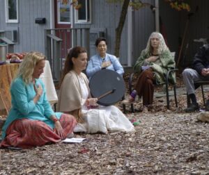 This is a photo of an outdoor cacao ceremony at Sanctuary Retreat Center. There are two female retreat leaders, one playing a handheld drum. There is a semi-circle of people around them.