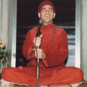 Master Charles Cannon wearing all orange with rudraksha beads and smiling while holding a microphone