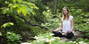 A woman sits cross-legged on a rock in a lush, green forest, meditating with her eyes closed and hands resting on her knees. Sunlight filters through the trees, casting light on her serene expression.