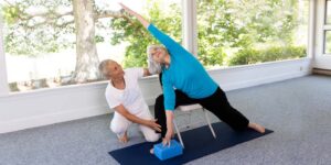 An older woman practicing a yoga pose using a chair and yoga block for support, guided by an instructor in a bright, spacious room with large windows overlooking trees.