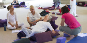 3 women in a yoga classroom studying a fourth woman in a reclined restorative yoga pose